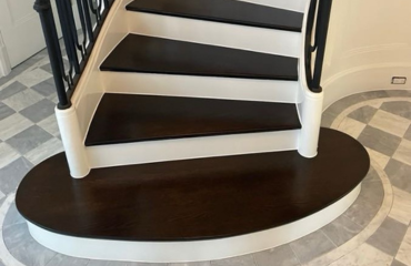 The image shows an elegant staircase with a combination of dark brown and white colors. The stair treads are dark brown, contrasting sharply with the white risers and stringers. The bottom step is curved, creating a half-moon shape that extends into the floor, which has a checkered pattern in light and dark shades, possibly a marble or tile finish.